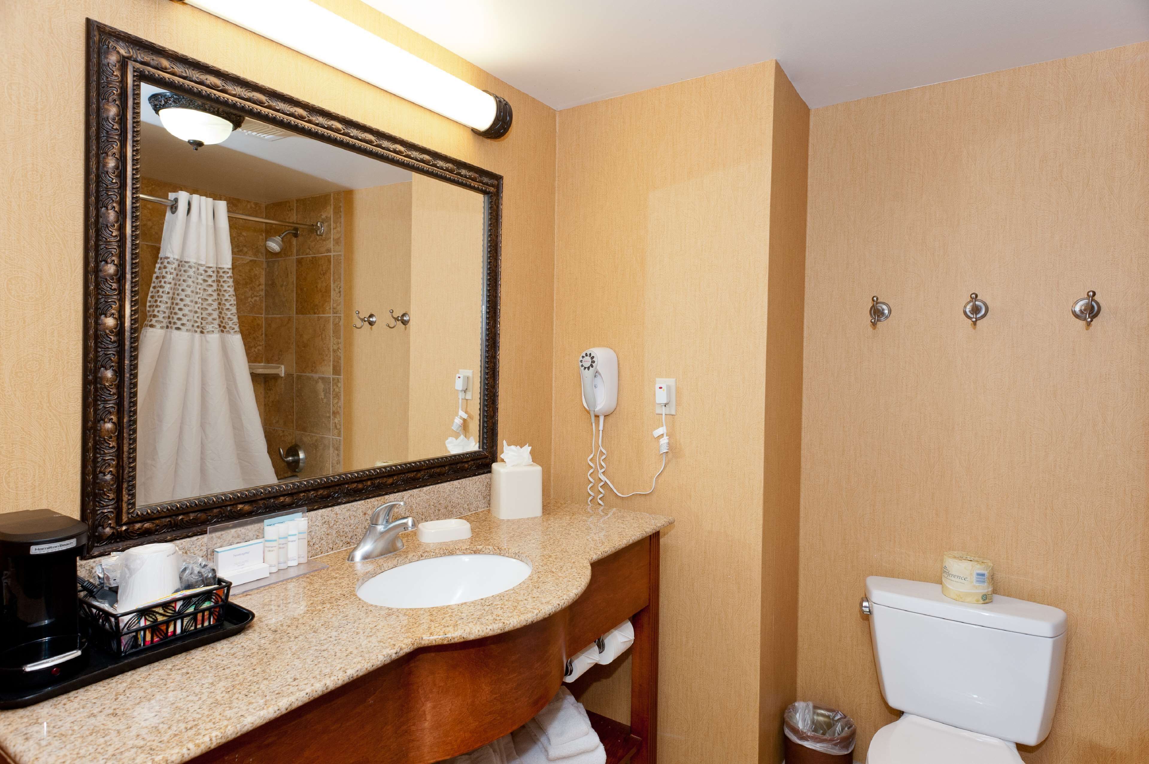A bathroom with a large mirror, sink, and toilet. There is a hairdryer and a trash can visible.
