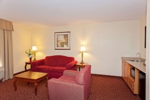 A hotel room with a red sofa, a pink armchair, a wooden coffee table, two lamps, a framed picture on the wall, and a kitchen area with a sink and cabinets.