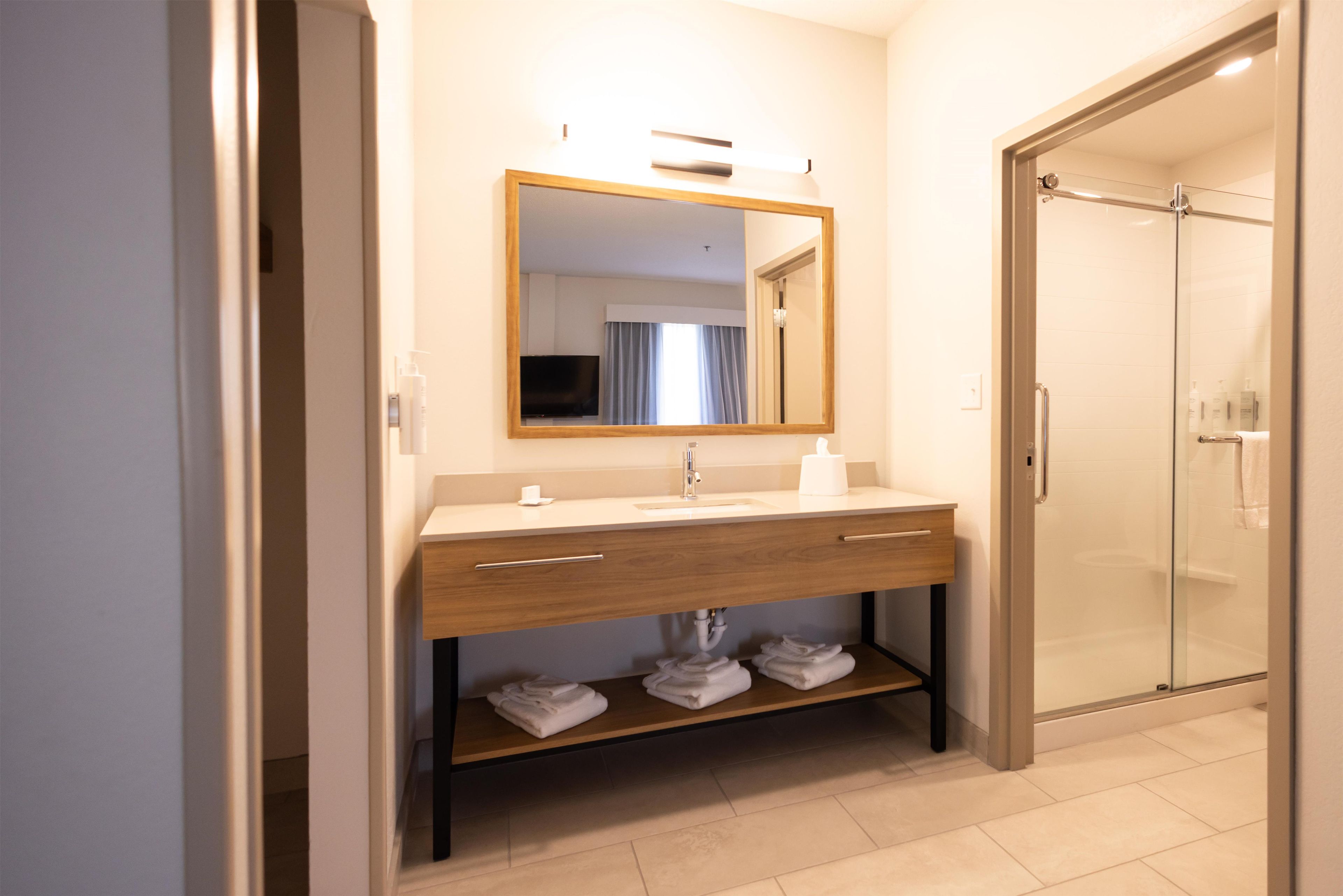 A modern bathroom with a wooden vanity, a mirror, a glass shower enclosure, and neatly folded towels on the floor.
