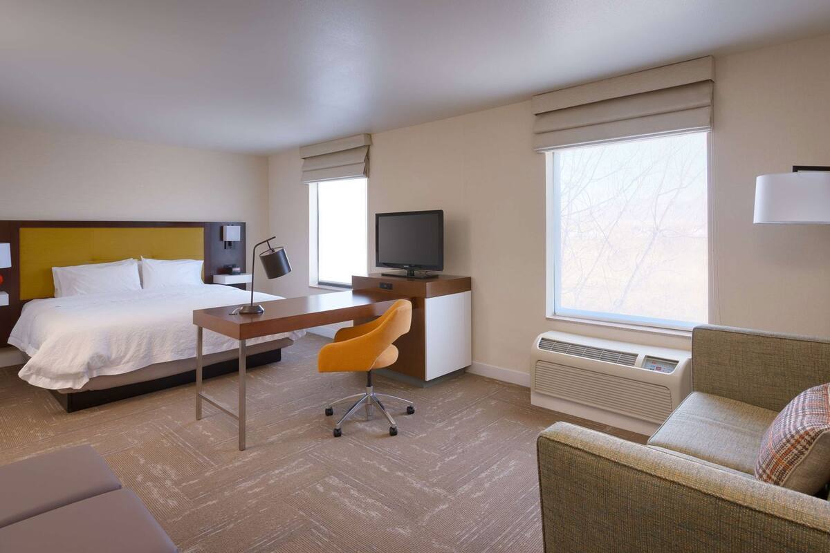 A hotel room with a bed, desk, chair, TV, and large windows.