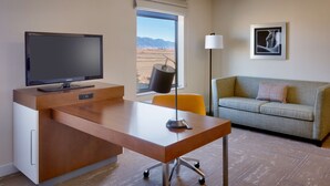 A hotel room with a wooden desk, a television, a sofa, and a window with a view of mountains.