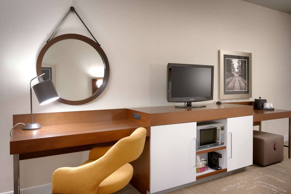 A modern hotel room with a wooden desk, a flat-screen TV, a microwave, a coffee maker, and a compact refrigerator.