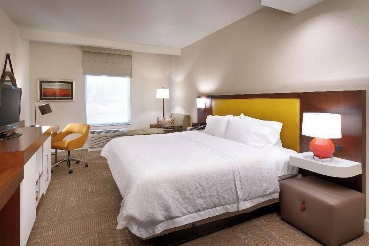 A hotel room with a bed, a desk with a computer, a chair, a nightstand, a lamp, and a window with curtains.