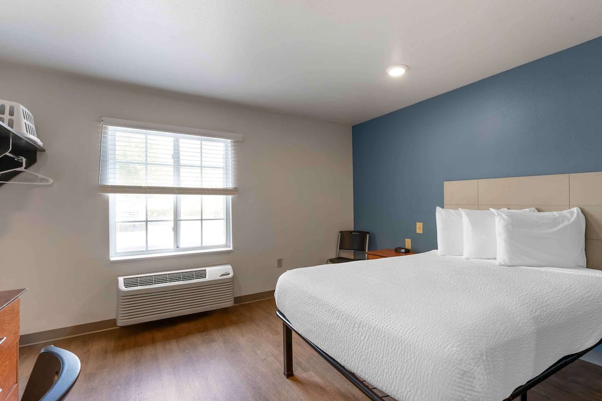 A bedroom with a large bed, a window with blinds, a heater, a nightstand, and a wall-mounted shelf.