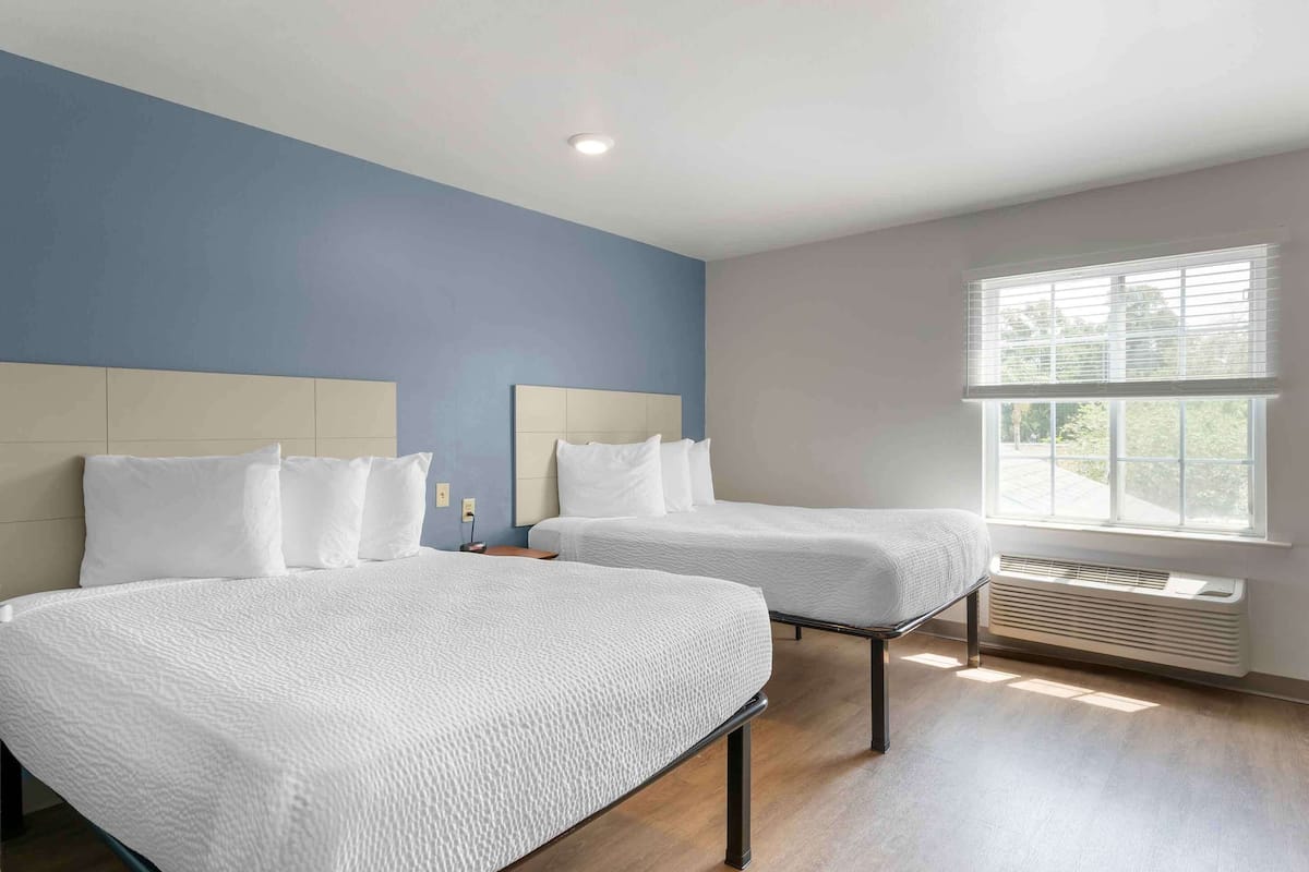 A hotel room with two beds, a window with blinds, a ceiling light, and a wall heater.