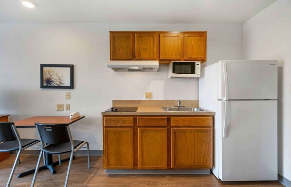 Una cocina compacta con armarios de madera, microondas, refrigerador, lavamanos y una zona de comedor con dos sillas y una mesa.