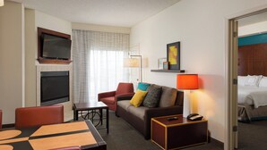 A hotel room with a flat-screen TV mounted above a fireplace, a sofa with colorful cushions, a dining table with chairs, and a bedside table with a lamp and phone. - Residence Inn by Marriott Kansas City Airport (Kansas City)
