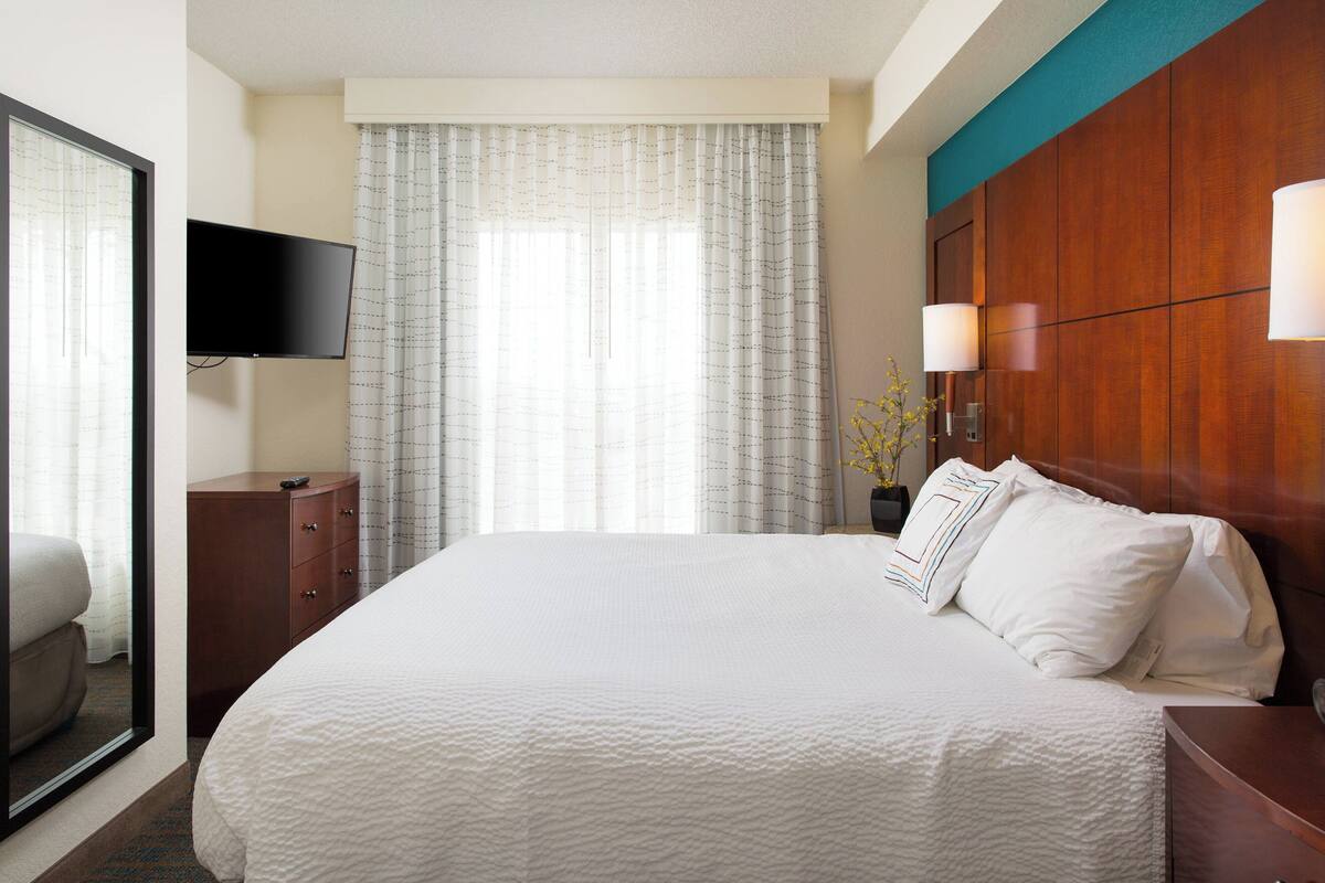 A hotel room with a large bed, a flat-screen TV mounted on the wall, a wooden headboard, and a window with sheer curtains.