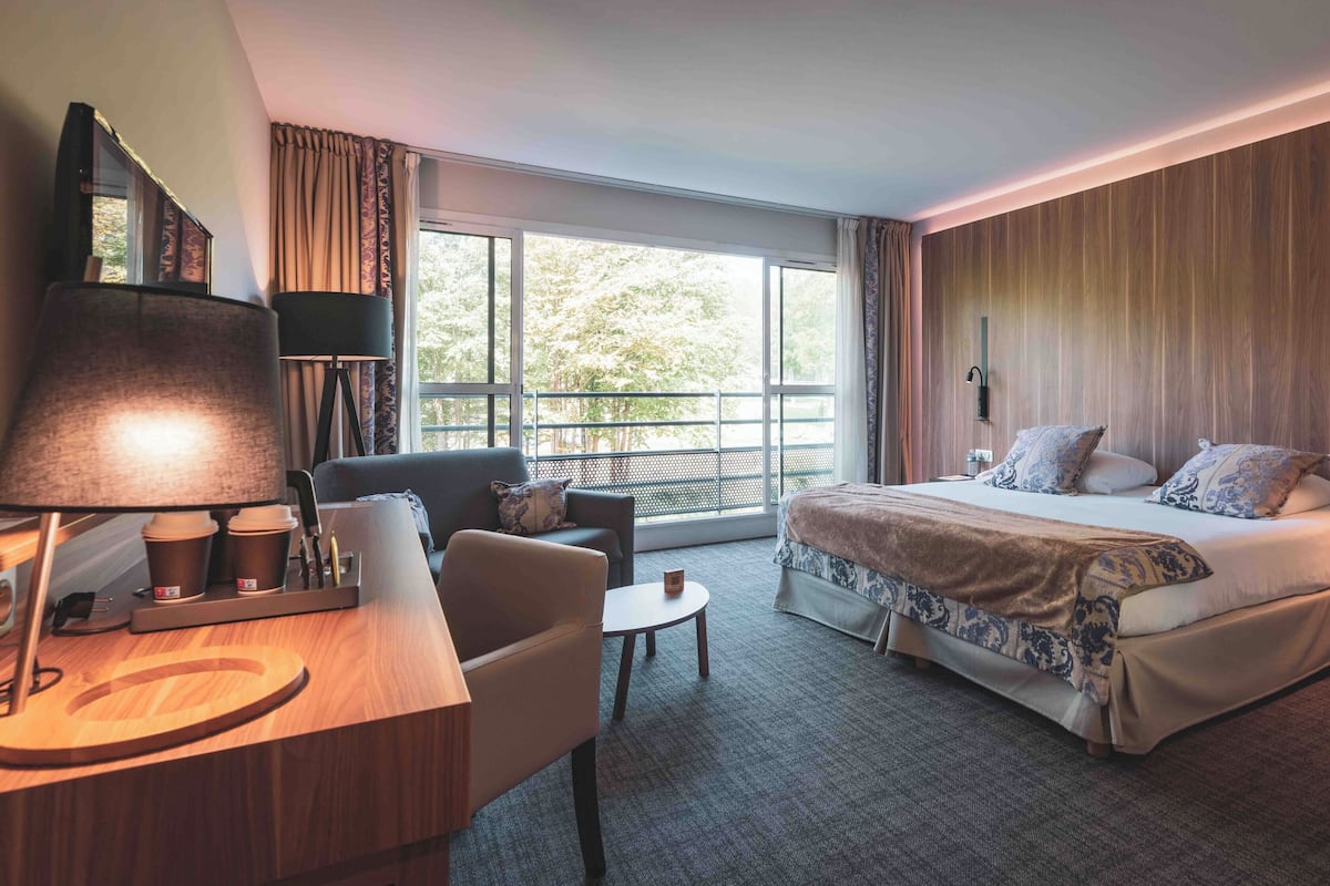 A hotel room with a bed, a desk with a lamp, a chair, and a view of the outdoors through large windows.