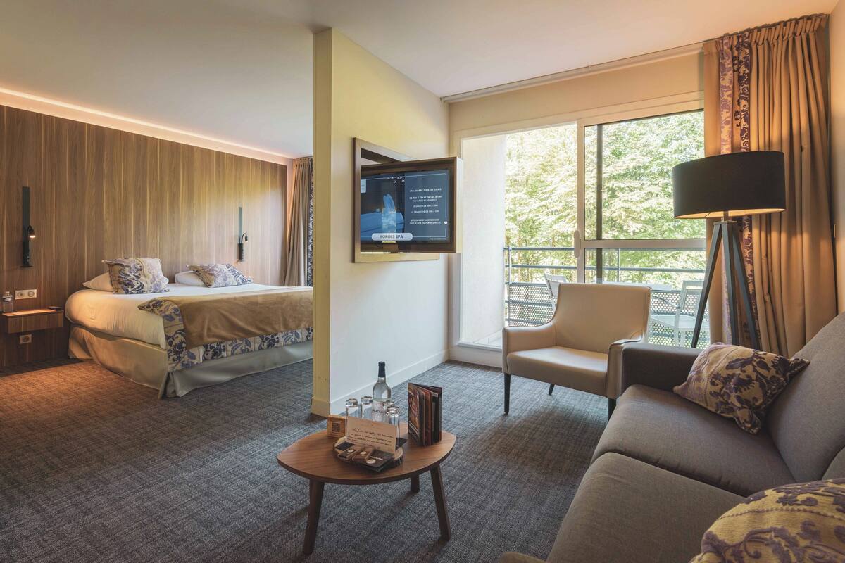 A hotel room with a bed, a sofa, a chair, a small table, and a TV mounted on the wall.