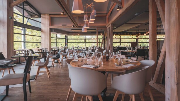 Un restaurante espacioso con vigas de madera, amplias ventanas e iluminación colgante moderna. Las mesas están preparadas con copas de vino y cubiertos.