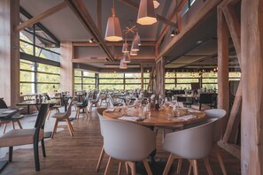 Um restaurante espaçoso com vigas de madeira, grandes janelas e iluminação pendente moderna. As mesas estão preparadas com copos de vinho e talheres.