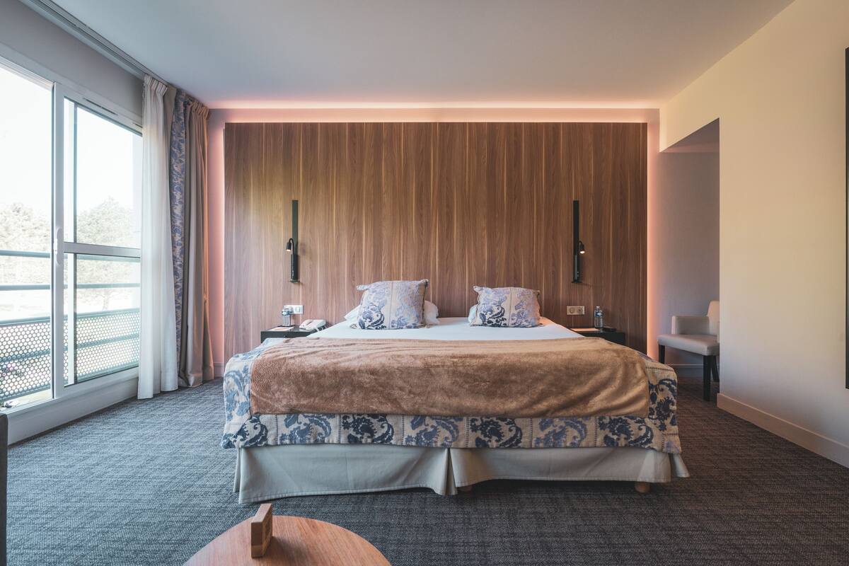 A modern hotel room with a large bed, a wooden accent wall, a view of the outdoors, and a small seating area.