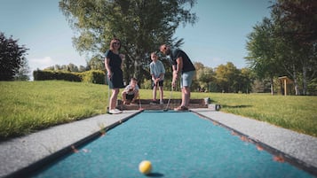 A croquet game in progress on a green field with a blue playing surface, surrounded by trees and grass.