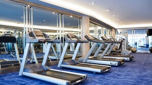 Varias cintas de correr en un gimnasio con alfombra azul y grandes ventanales al fondo.