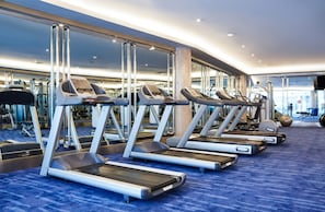 Une rangée de tapis de course dans une salle de sport, avec un tapis bleu et de grandes fenêtres en arrière-plan.