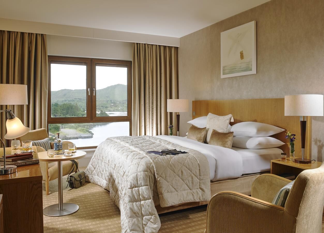 A hotel room with a large bed, two bedside tables with lamps, a desk with a chair, and a view of the outdoors through a large window.