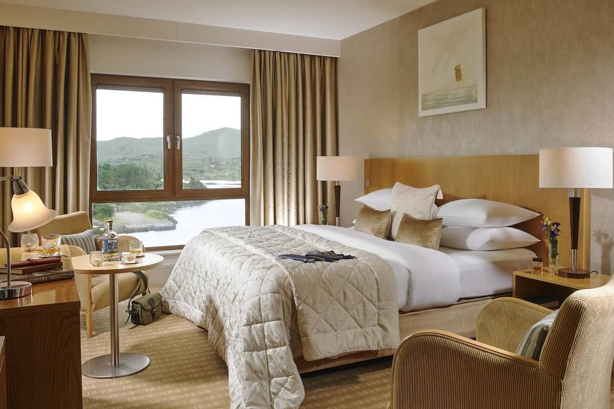 A hotel room with a large bed, two bedside tables with lamps, a desk with a chair, and a view of the outdoors through a large window.