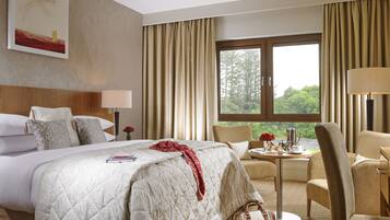 A hotel room with a large bed, a bedside table with a lamp, a chair, a small table with a vase of flowers, and a window with curtains.