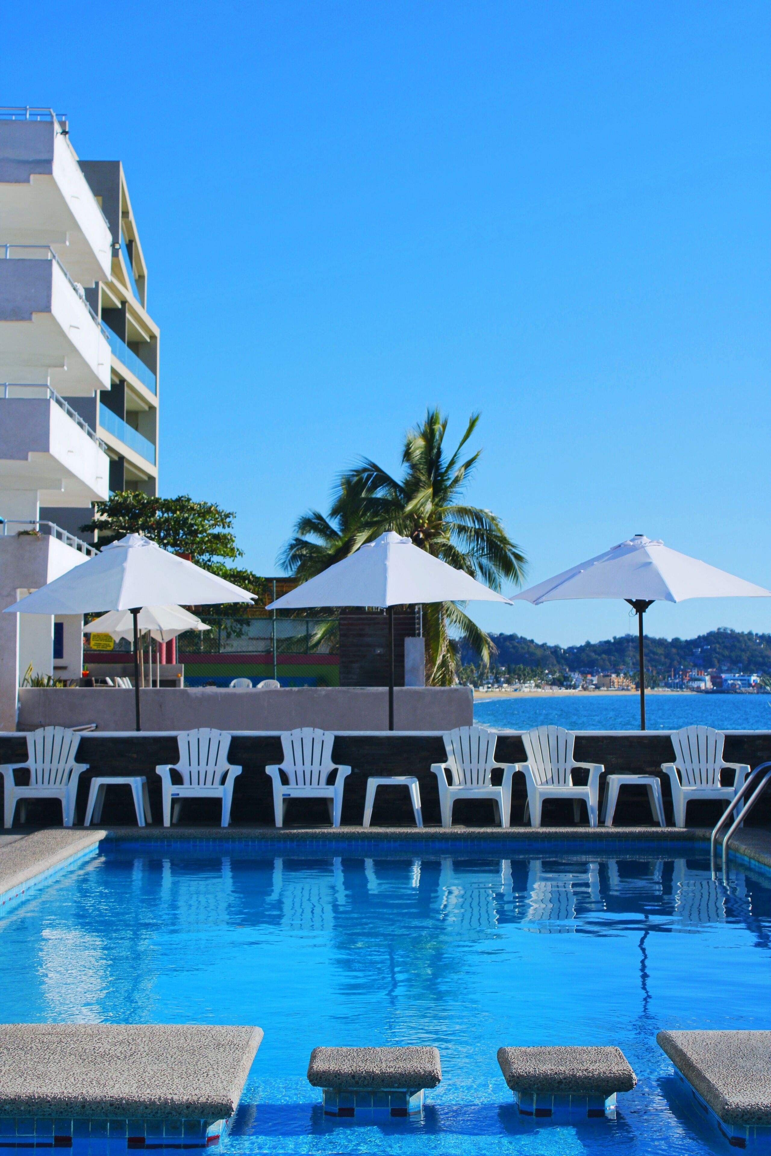 A swimming pool with white chairs and umbrellas, a building with balconies, and a view of the sea and hills.