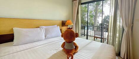A hotel room with a large bed, a plush toy on the bed, and a balcony with a view of trees.