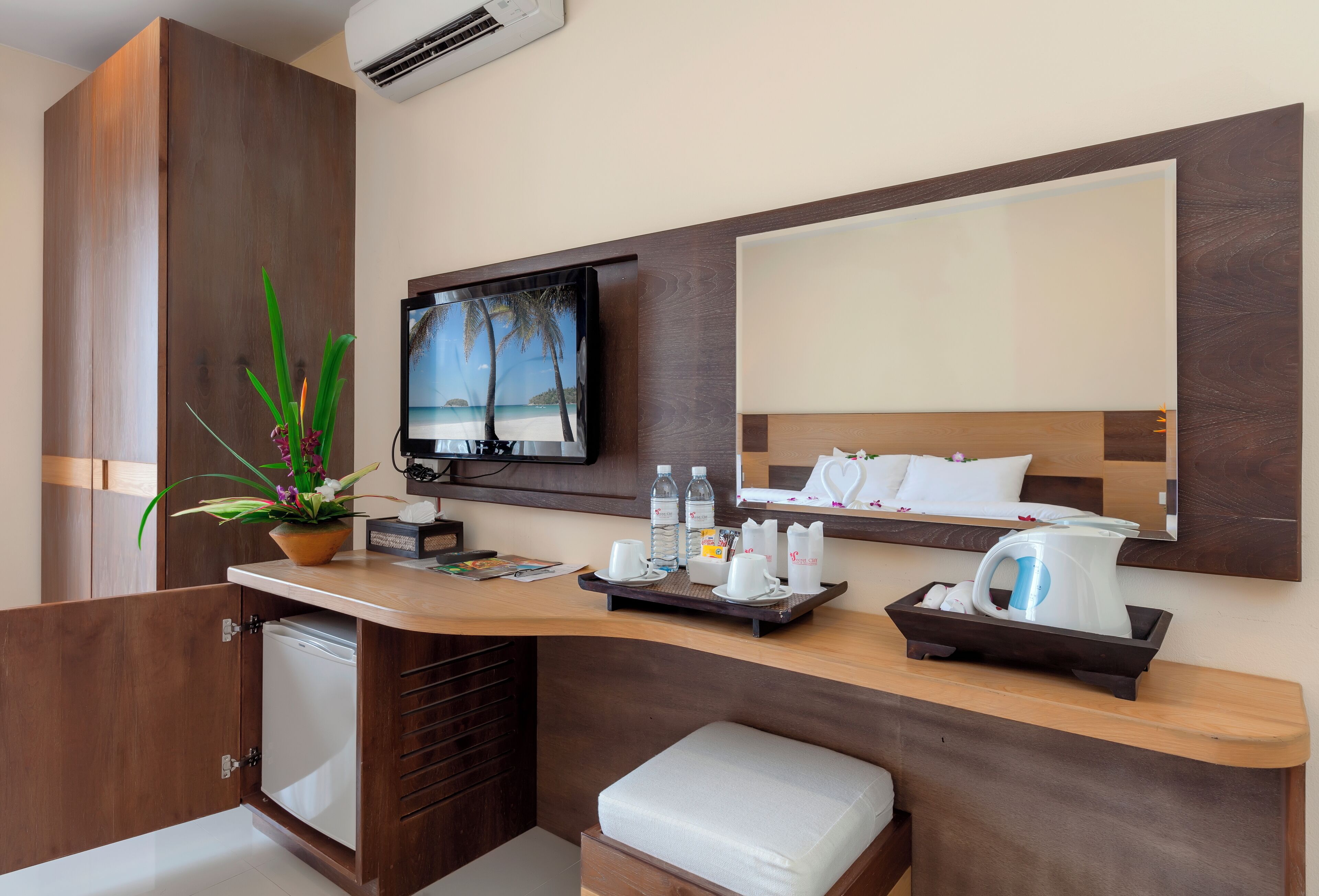 A modern hotel room with a wooden desk, a flat-screen TV, a small refrigerator, a coffee maker, and a potted plant.