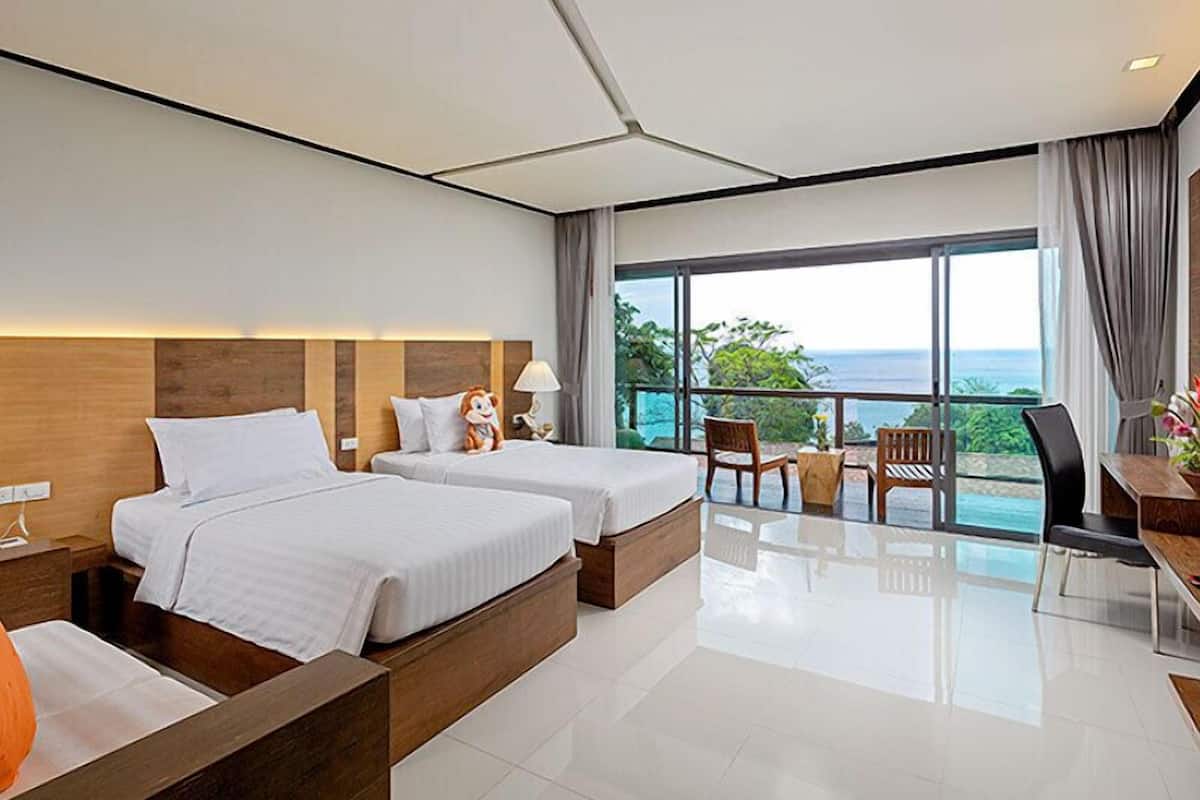 A modern hotel room with two beds, a balcony with a view of the ocean, and a desk area.