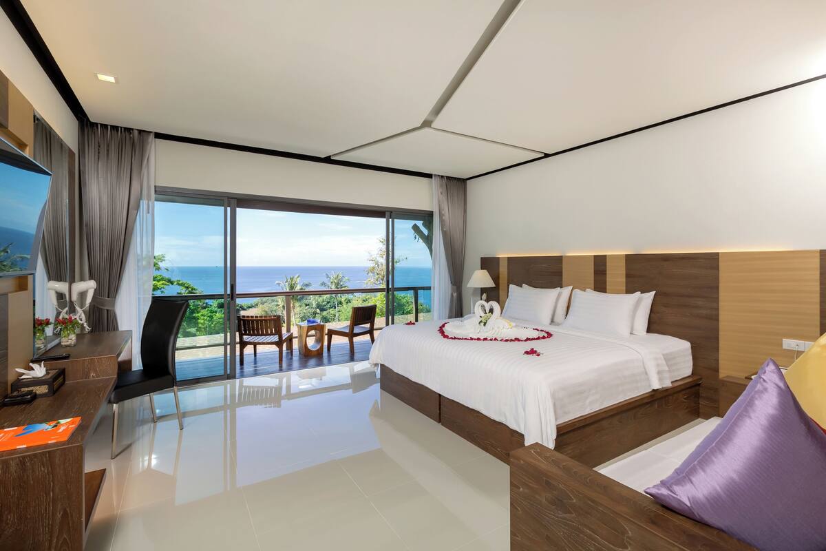 A modern hotel room with a large bed, a desk, and a balcony with a view of the ocean.