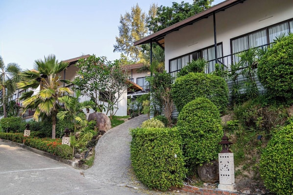 Un edificio moderno con fachada blanca, ventanas grandes y techo inclinado. Abundan plantas y árboles verdes alrededor de la propiedad.