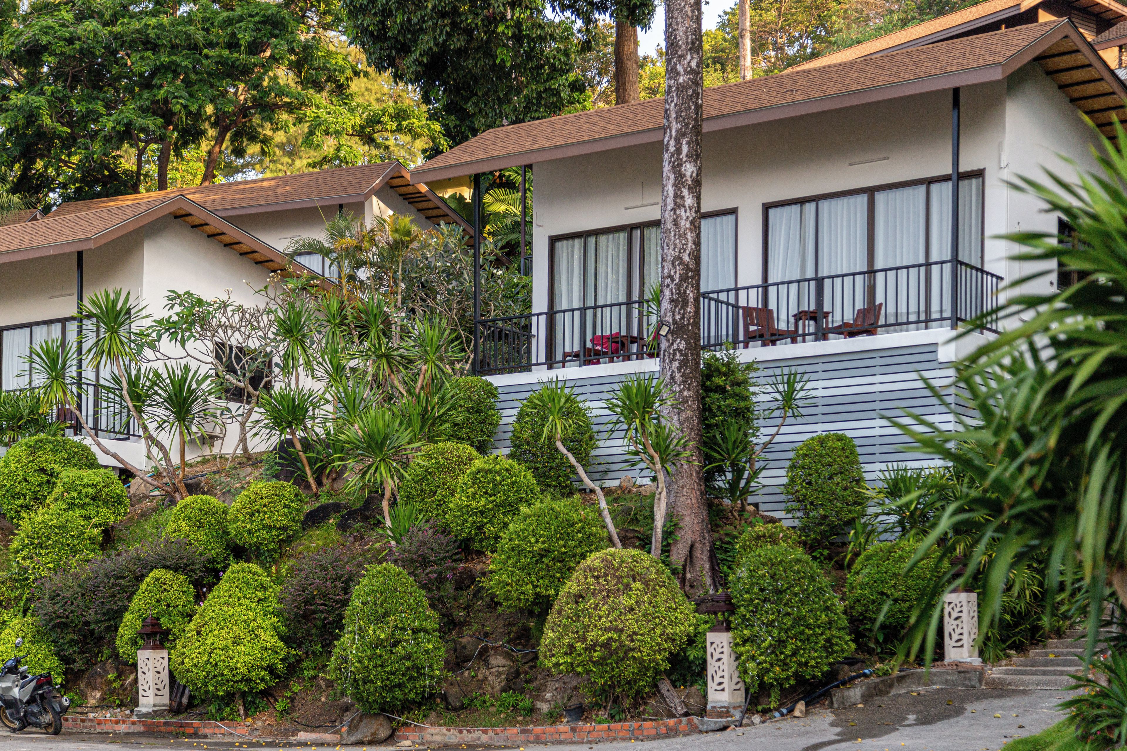 A hotel with multiple-story buildings, a balcony with seating, and a well-maintained garden with various shrubs and trees.