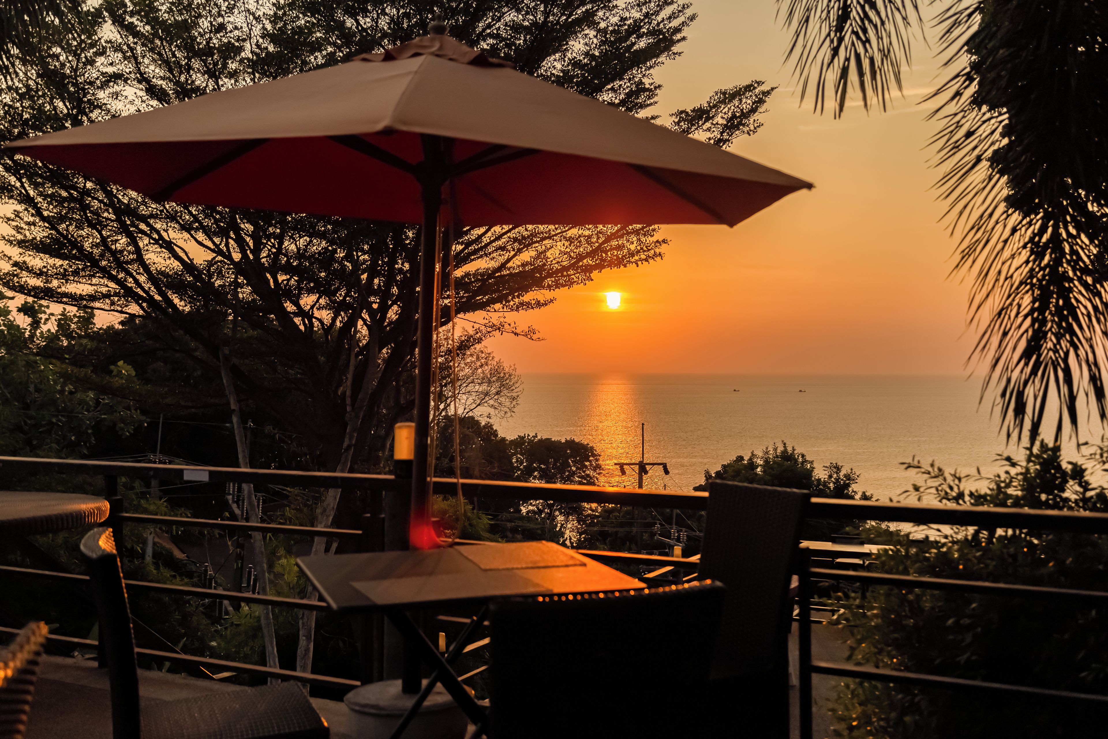 An outdoor seating area with a table and an umbrella, overlooking a sunset view of the ocean.