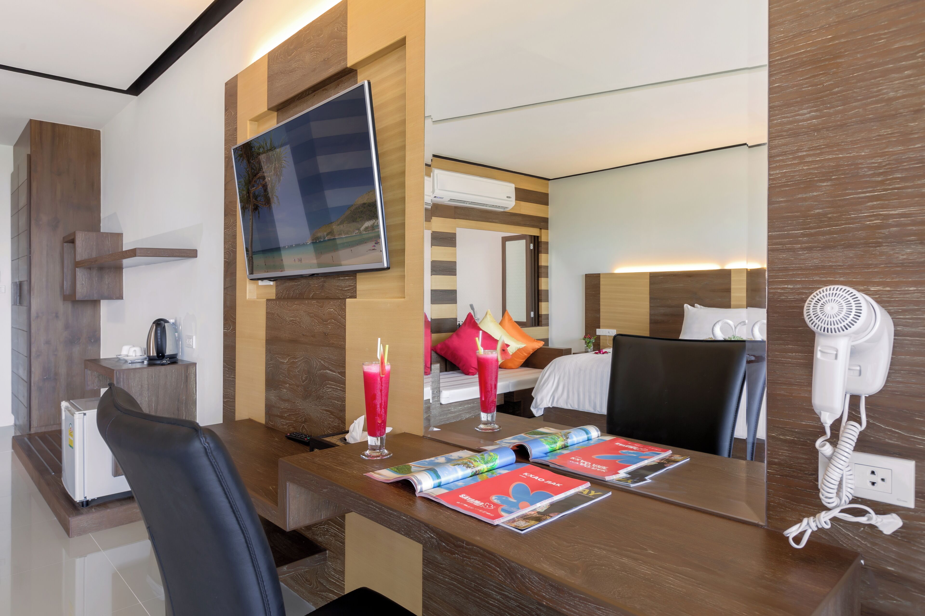 A hotel room with a wooden desk, a flat-screen TV, a bed with pillows, a hairdryer, and a mirror.