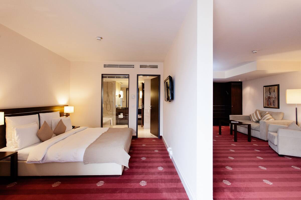 A hotel room with a bed, a desk, a chair, a sofa, a TV, and a bathroom visible through an open door.