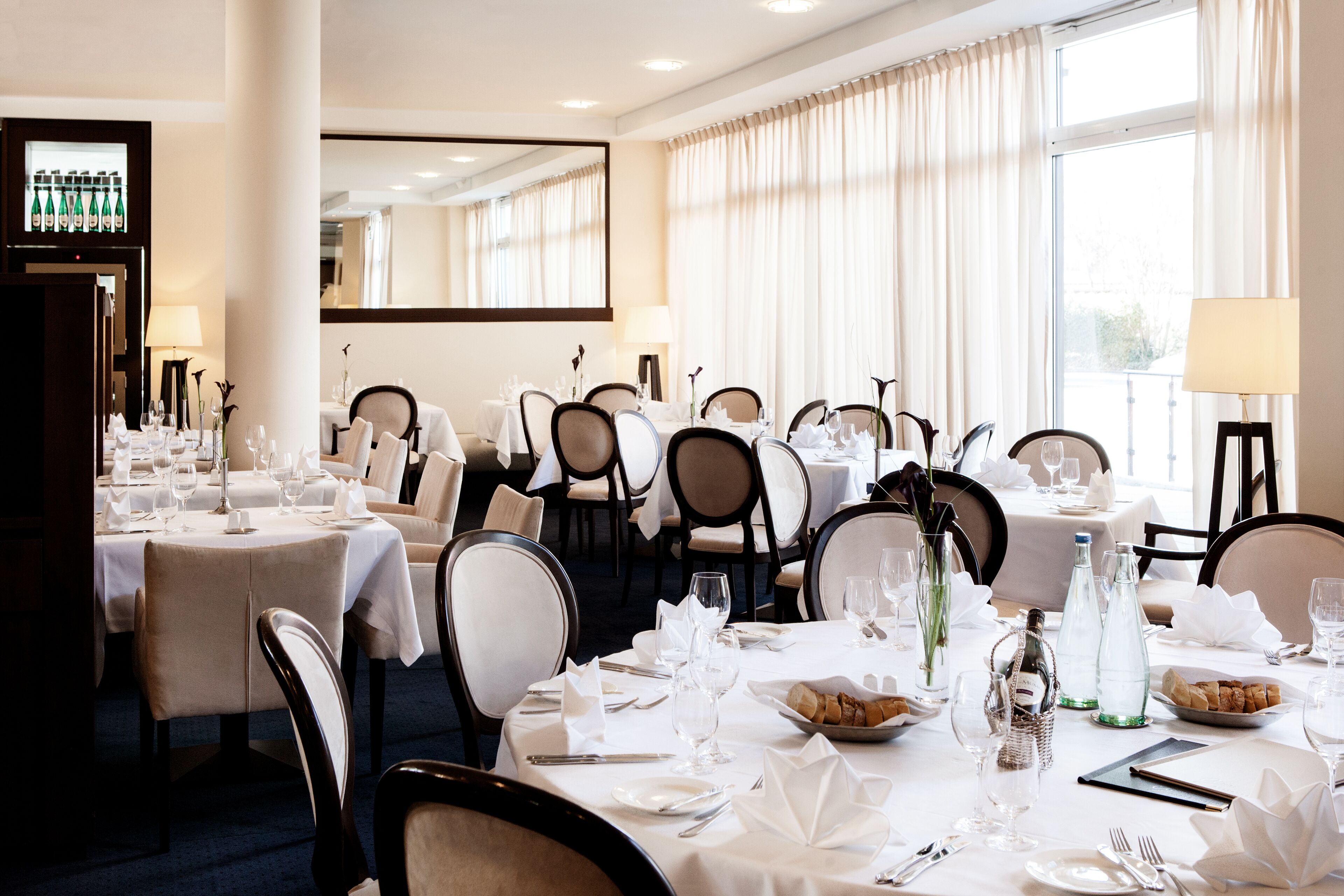 A hotel dining area with multiple tables set for dining, each with chairs, white tablecloths, and various dining utensils.