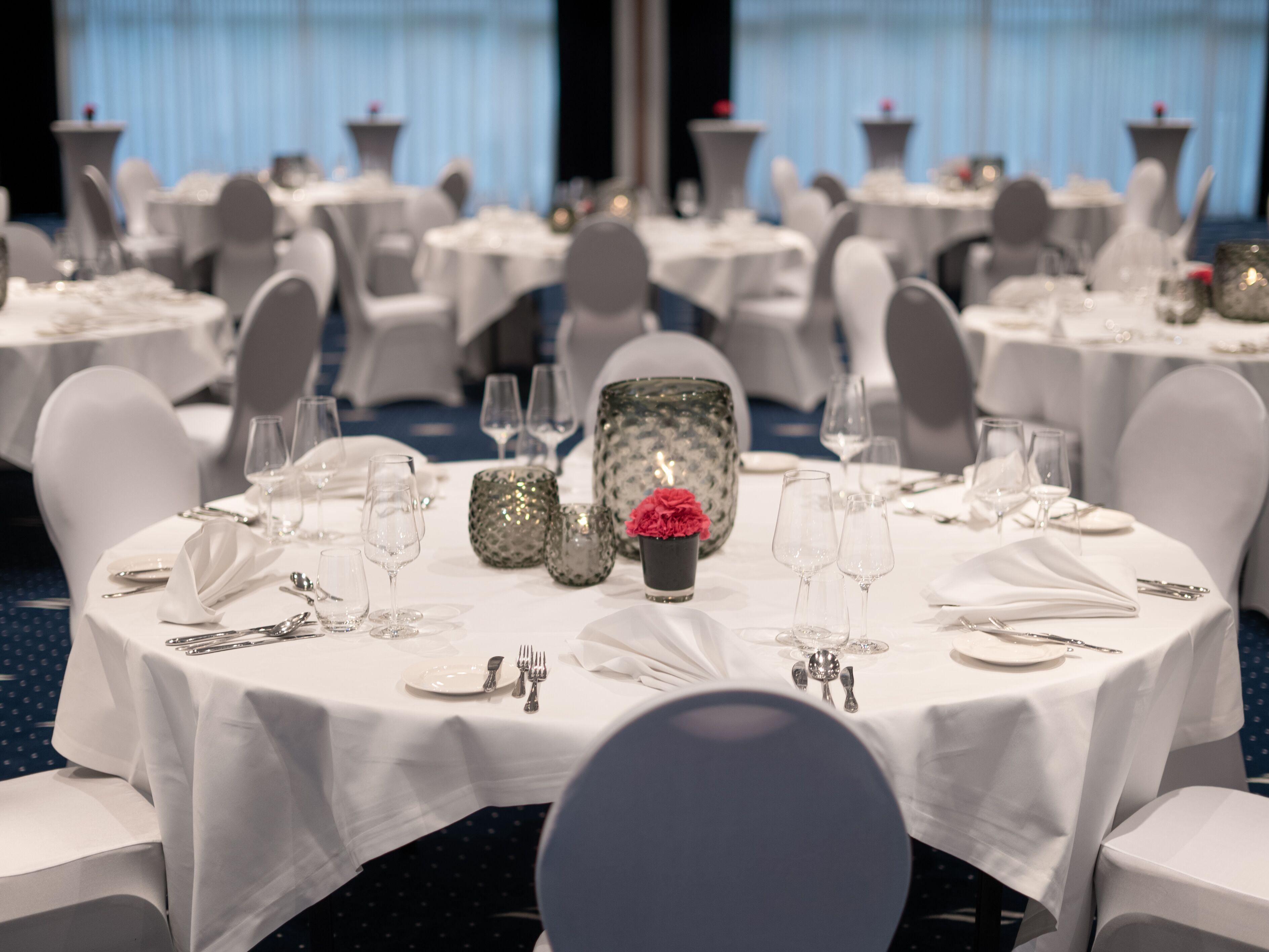 Un salón de banquetes con mesas redondas dispuestas para comer, cada una con mantel blanco, cubiertos y cristalería.