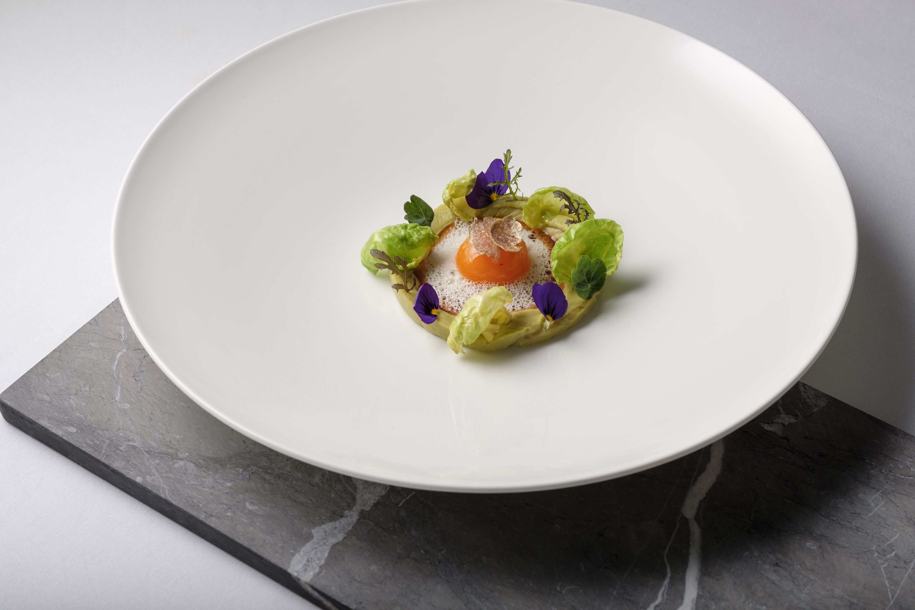 A white plate with a central egg yolk surrounded by greens and purple flowers on a marble surface.