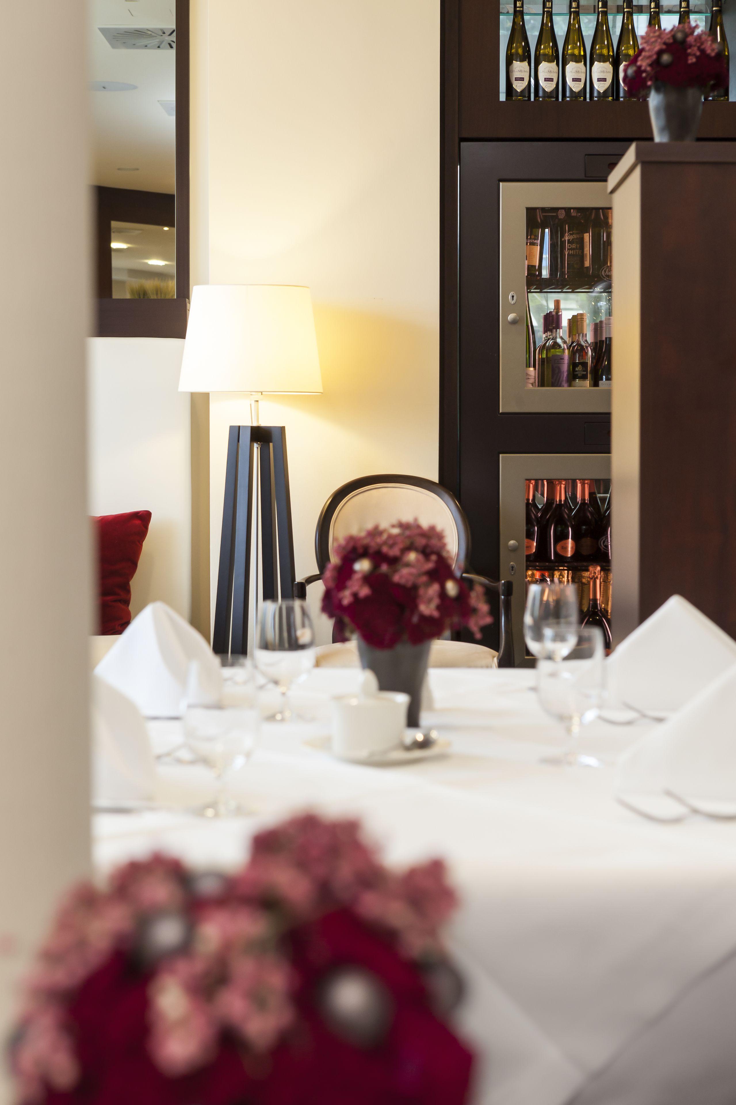 A dining table set with white tablecloth, a vase of flowers, and a wine cooler in the background.