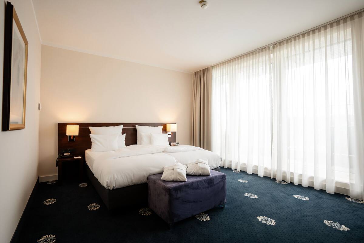 A hotel room with a large bed, a nightstand, a lamp, a window with sheer curtains, and a dark carpet with a floral pattern.