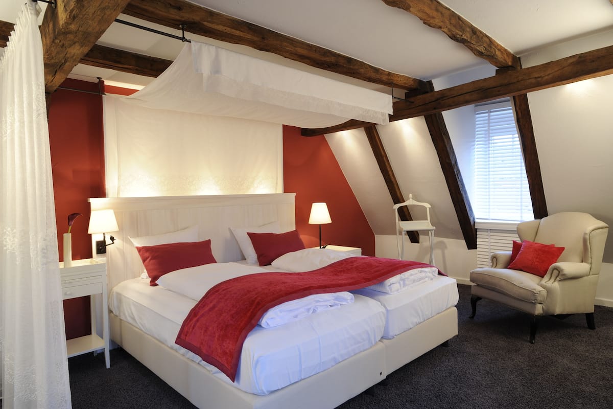A bedroom with a four-poster bed, a chair, a nightstand, and a window with blinds.