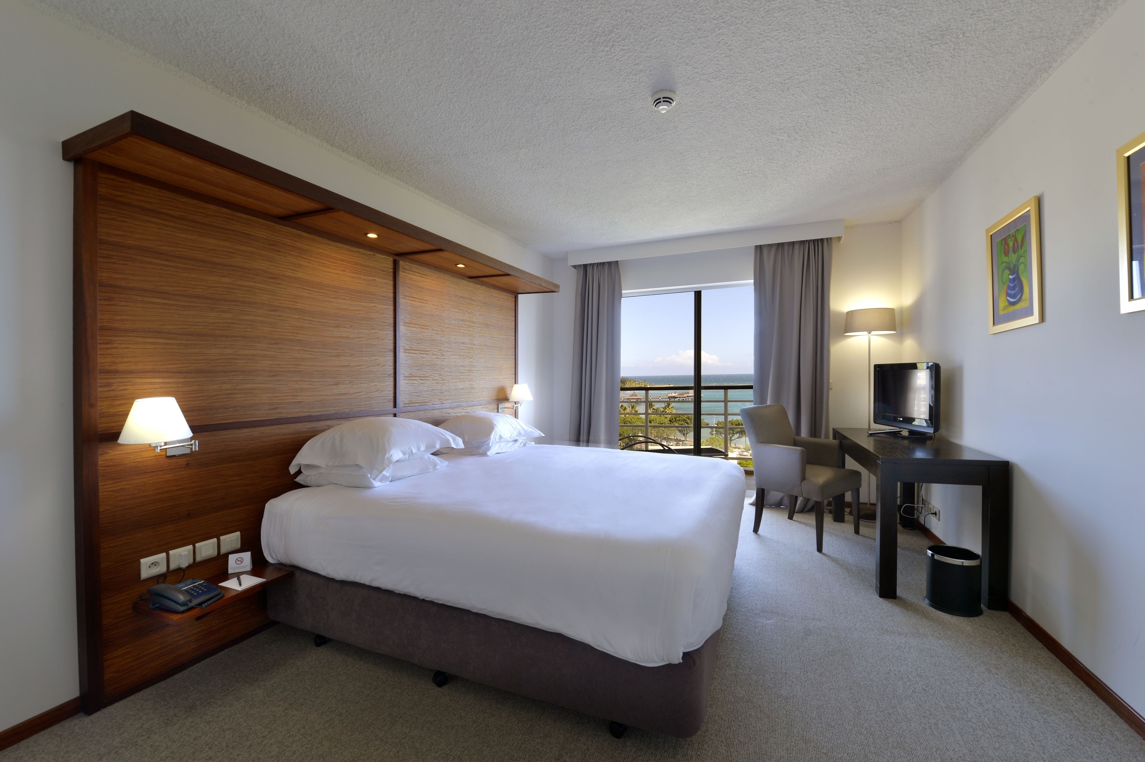 Ein Hotelzimmer mit einem großen Bett, Nachttisch mit Lampe, Fernseher und Blick ins Freie.