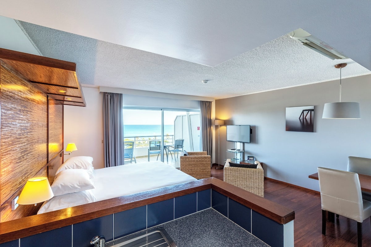 A hotel room with a large bed, a desk, a chair, a TV, and a view of the ocean.