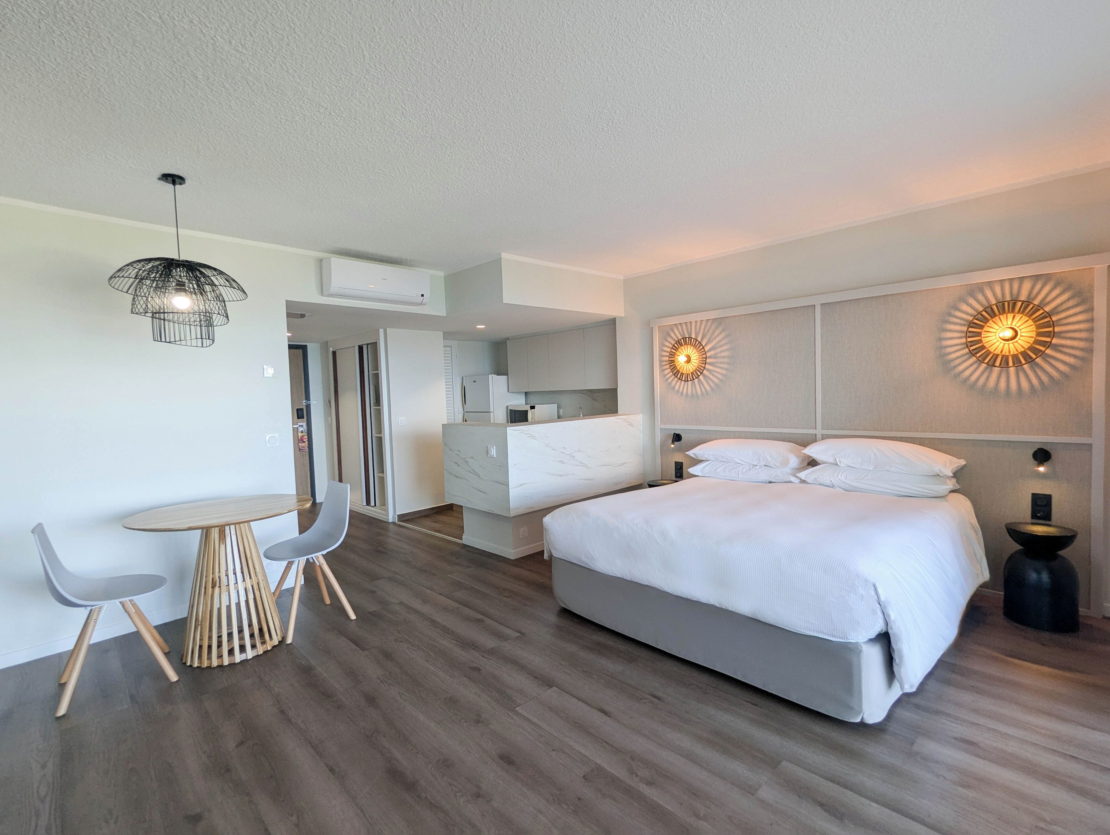 A modern hotel room with a large bed, a dining area with a table and chairs, a kitchen area, and a bathroom.
