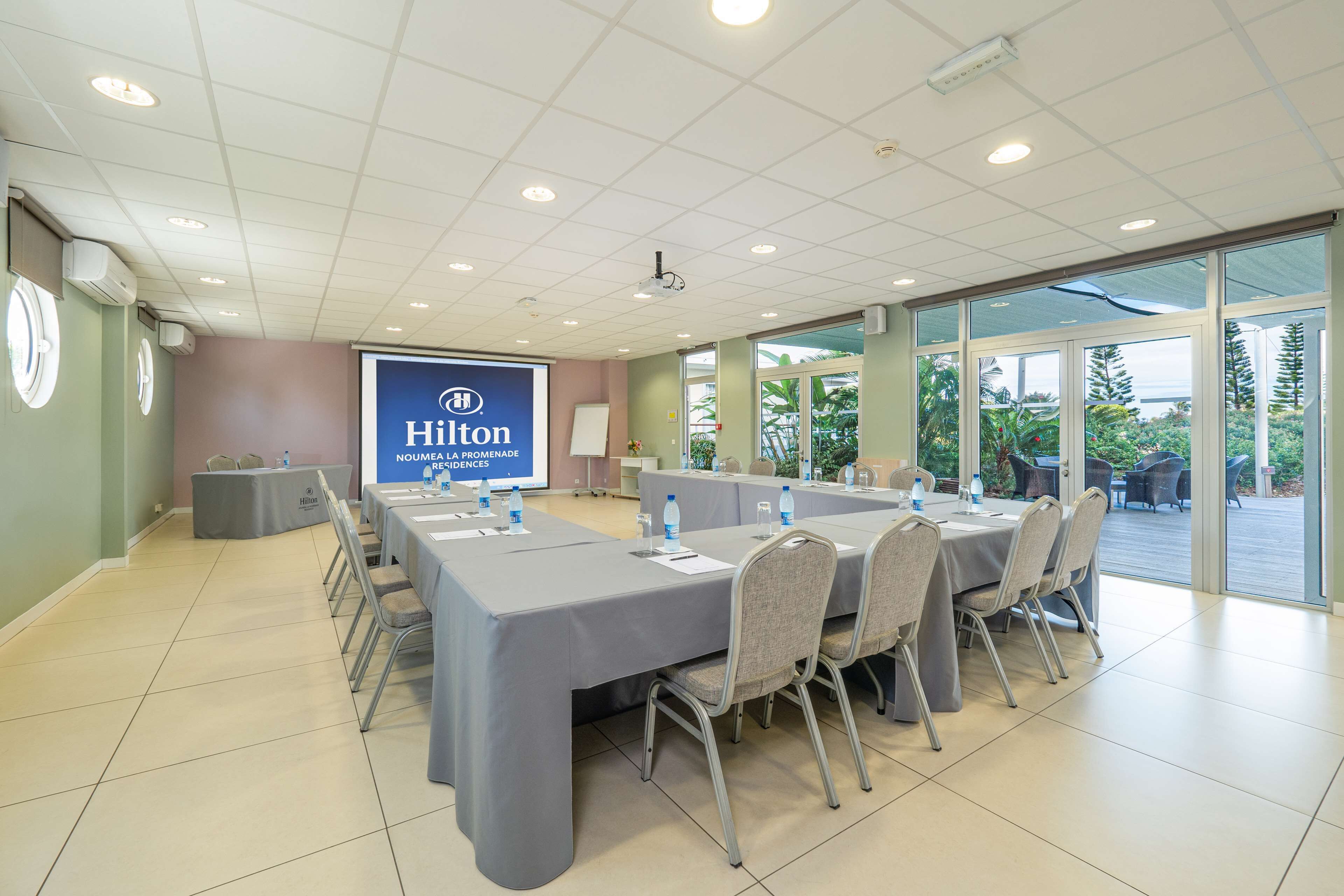 Un salón de conferencias con una larga mesa, sillas, una pantalla de proyector y vistas al exterior.