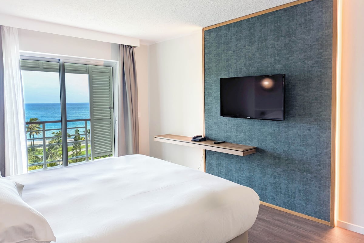 A hotel room with a bed, a TV mounted on a textured wall, a small wooden shelf, and a view of the ocean through a large window with curtains.