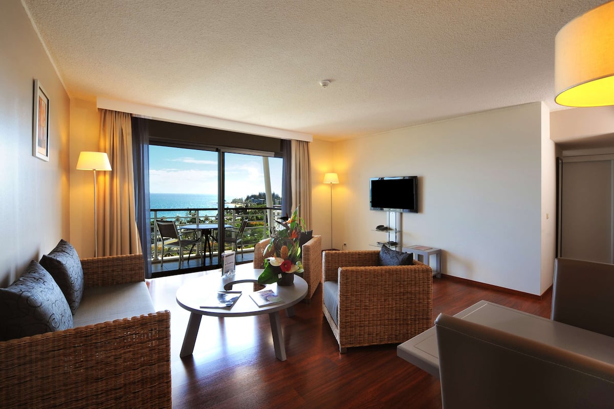 A modern hotel room with a balcony, wicker furniture, a flat-screen TV, and a view of the ocean.