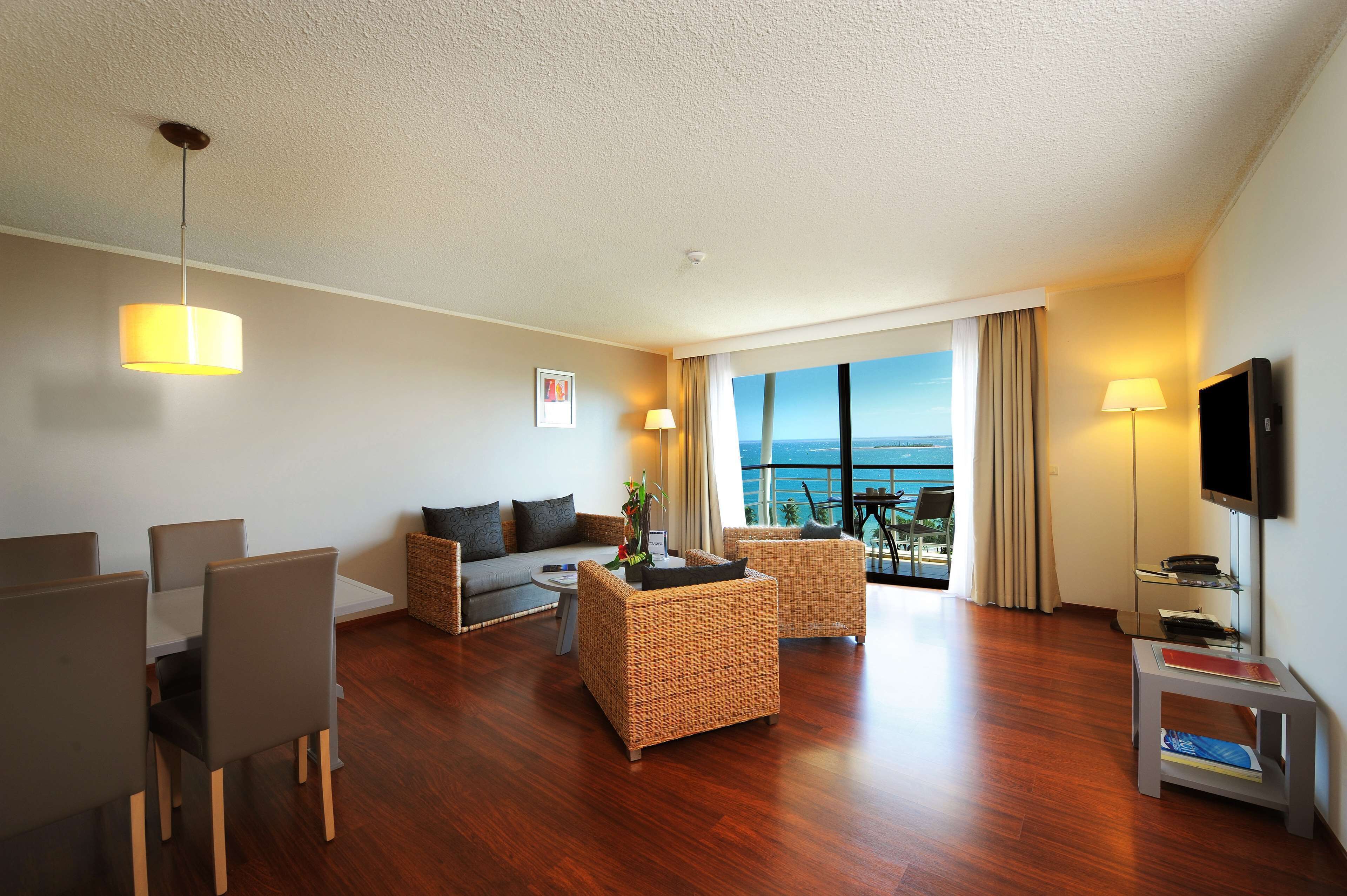A modern hotel room with a dining area, a sofa, two armchairs, a flat-screen TV, and a large window offering a view of the sea.