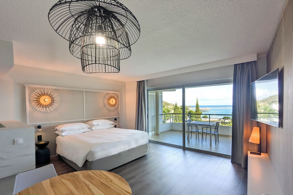 A modern hotel room with a large bed, a wooden table, and a balcony with a view of the sea.