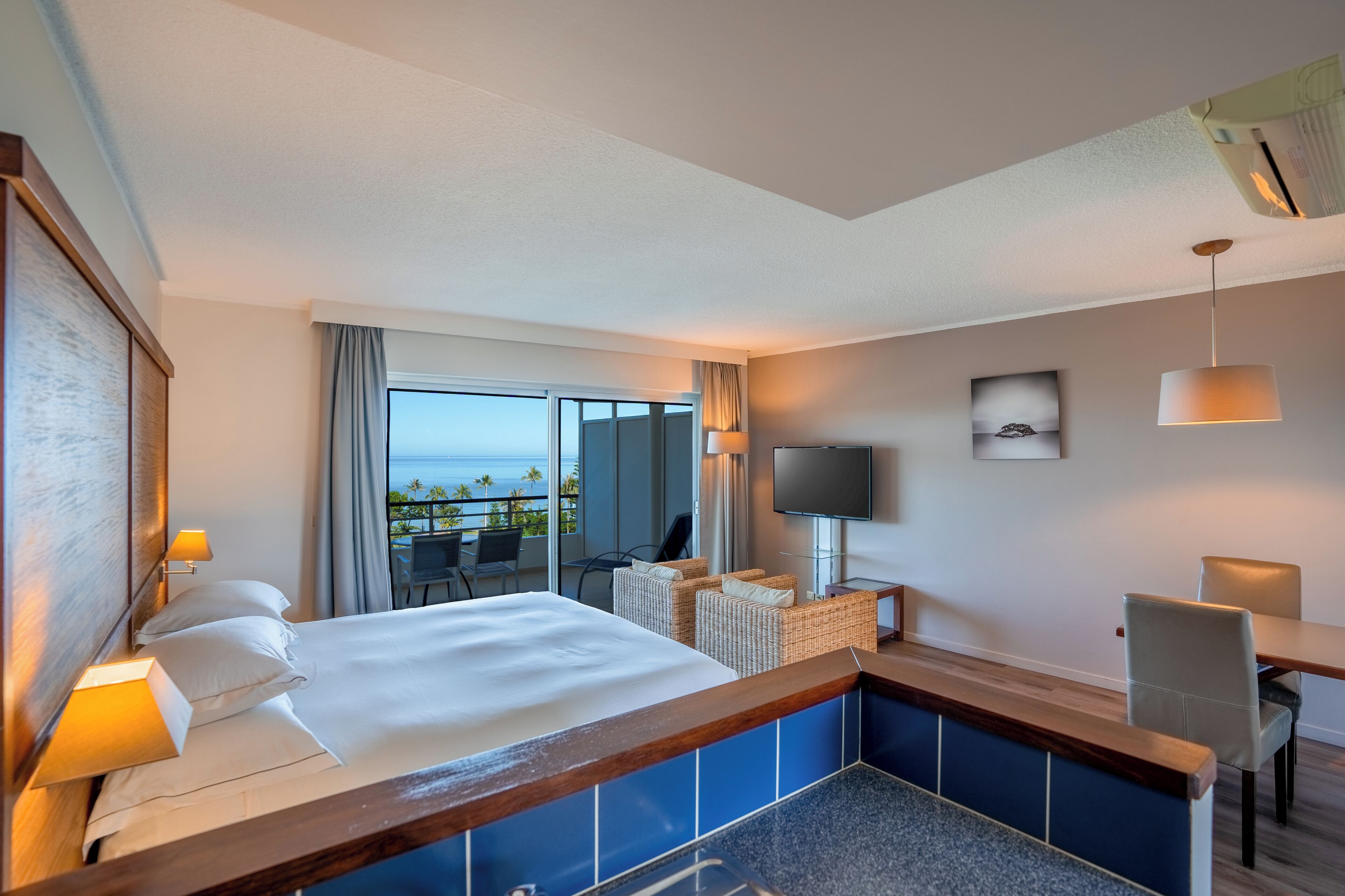 A hotel room with a large bed, a TV, a desk, and a view of the ocean.