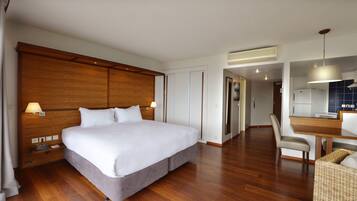 A hotel room with a large bed, wooden headboard, bedside tables with lamps, a dining area with a table and chairs, and a kitchenette.