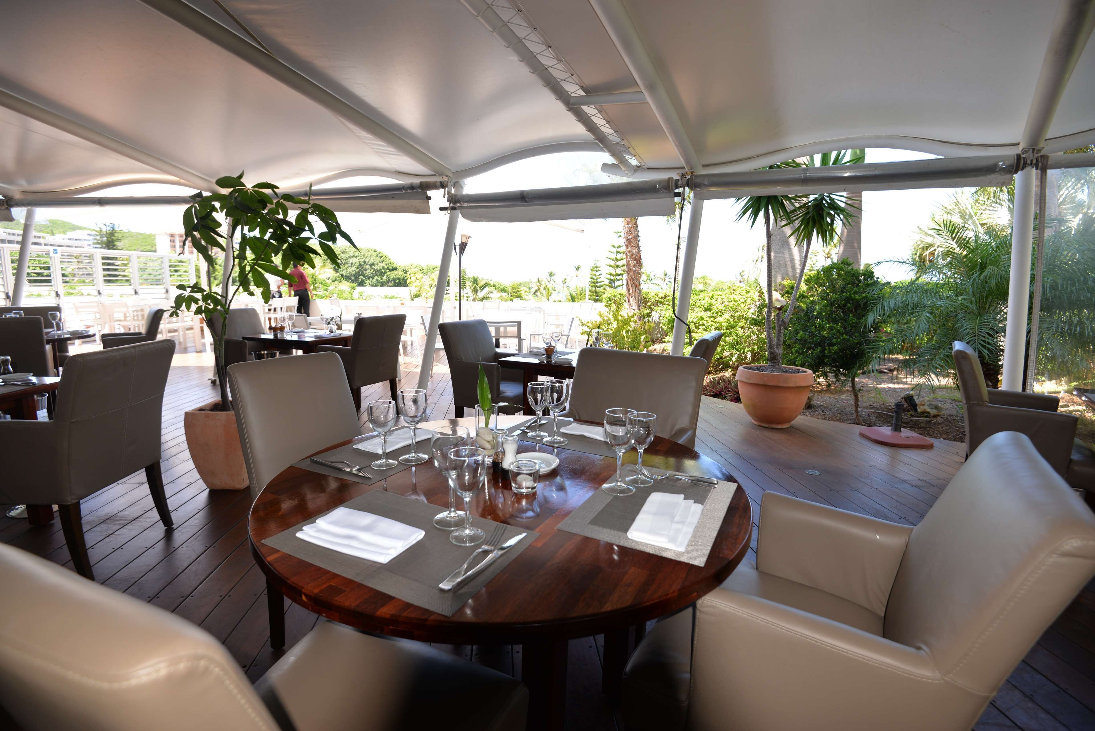 Un espace extérieur aménagé pour les repas, avec des tables rondes, des chaises blanches dotées de coussins et une grande plante en pot.